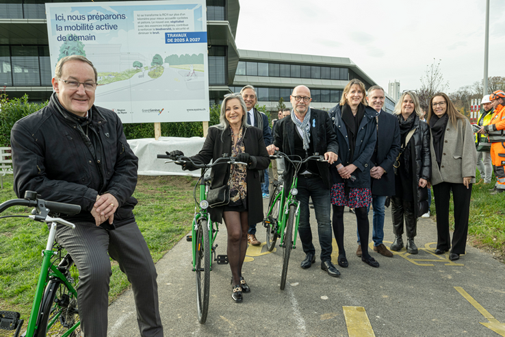 les personnes posent sur une route, appuyée sur des vélos. Derrière, au bord de la chaussée, un panneau de chantier. On lit: "Ici nous préparons la mobilité active de demain." On voit deux ouvriers casqués.