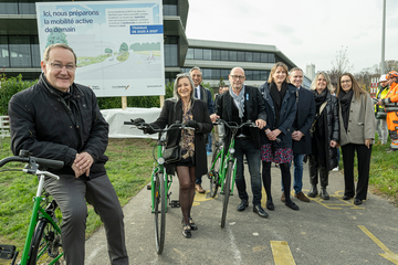 les personnes posent sur une route, appuyée sur des vélos. Derrière, au bord de la chaussée, un panneau de chantier. On lit: "Ici nous préparons la mobilité active de demain." On voit deux ouvriers casqués.