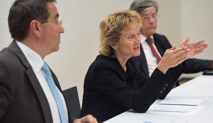 Les deux personnages assis à une table. Mme Widmer-Schlumpf parle.