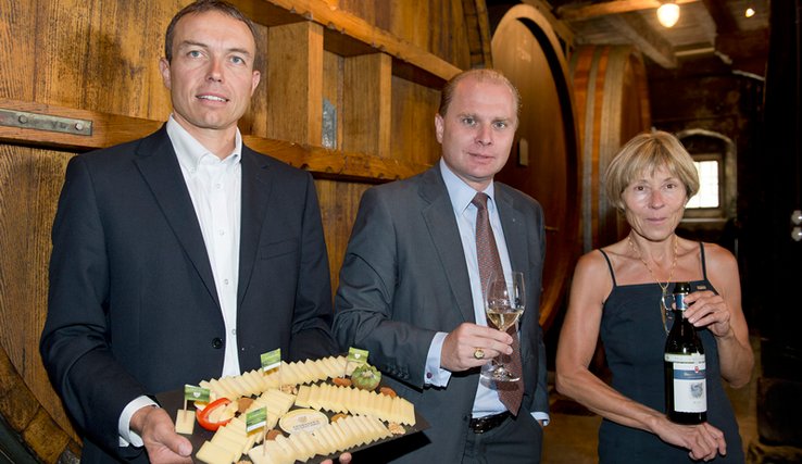 René Pernet, maître fromager, tient un plateau de fromage. Binia Ris de Fischer, propriétaire du domaine, présente une bouteille de vin. Les deux personnages entourent le conseiller d'Etat Philippe Leuba, qui tient un verre de vin.