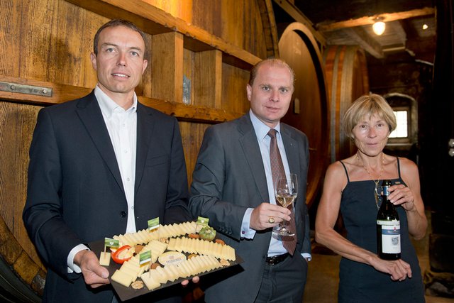 René Pernet, maître fromager, tient un plateau de fromage. Binia Ris de Fischer, propriétaire du domaine, présente une bouteille de vin. Les deux personnages entourent le conseiller d'Etat Philippe Leuba, qui tient un verre de vin.