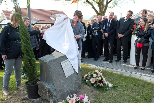 Une femme dévoile une stèle entourée de fleurs et d'un thuya, avec une plaquette au nom de M. Mermoud. A l'arrière plan, plusieurs membres du Conseil d'Etat.