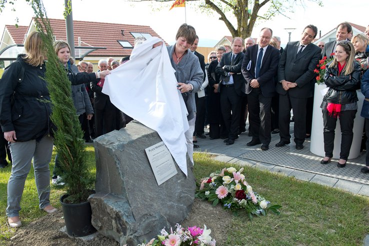 Une femme dévoile une stèle entourée de fleurs et d'un thuya, avec une plaquette au nom de M. Mermoud. A l'arrière plan, plusieurs membres du Conseil d'Etat.
