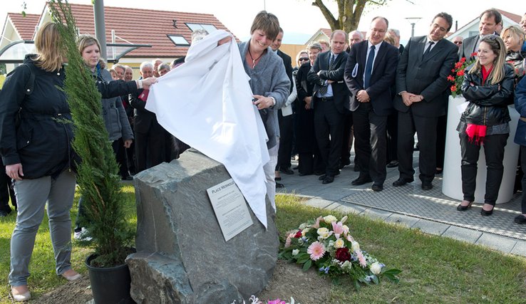 Une femme dévoile une stèle entourée de fleurs et d'un thuya, avec une plaquette au nom de M. Mermoud. A l'arrière plan, plusieurs membres du Conseil d'Etat.