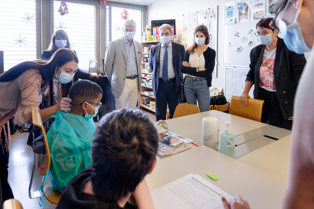 Les personnes sont debout dans une salle de classe, autour d'une table où sont assis deux enfants dont l'un porte une blouse. Tout le monde porte un masque sanitaire.
