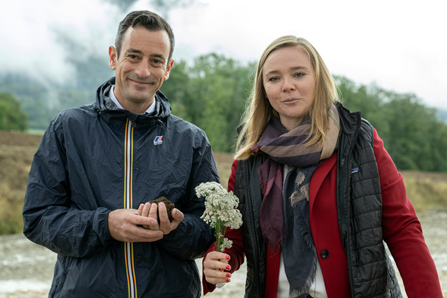 Les deux personnages posent dans une carrière près des Clées. Monsieur Venizelos tient une poignée de terre dans le creux de sa main, et madame Dittli, une fleur.