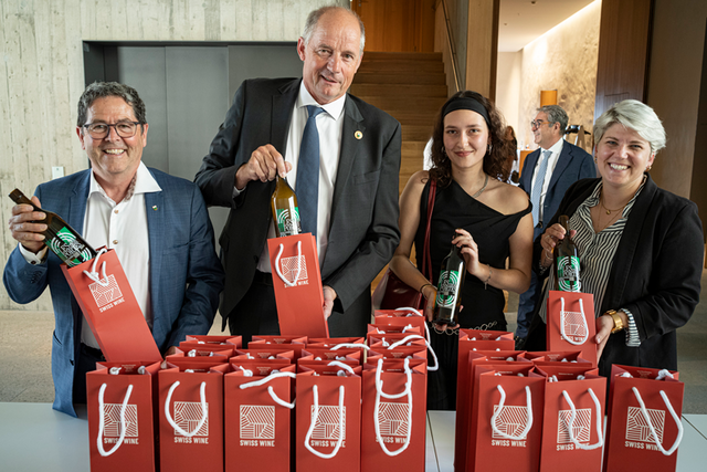 Les personnages posent derrière une table où sont déposées de nombreuses bouteilles emballées dans des sachets-cadeaux.