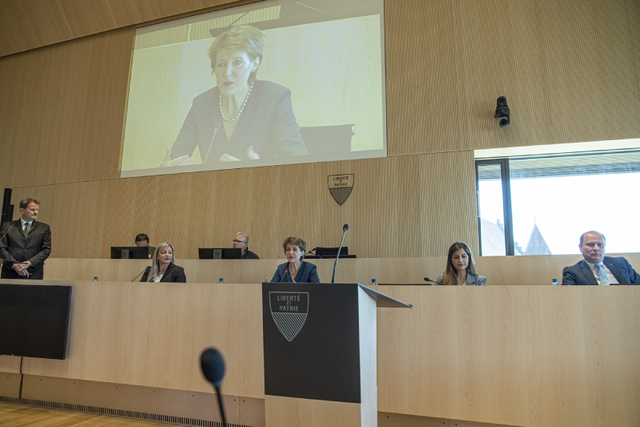 Mme Sommaruga est à la tribune de la salle du Grand Conseil. Son visage sur l'écran géant au-dessus. Les membres du Conseil d'Etat sont assis de part et d'autre.