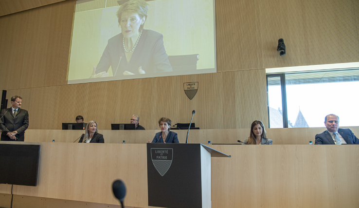 Mme Sommaruga est à la tribune de la salle du Grand Conseil. Son visage sur l'écran géant au-dessus. Les membres du Conseil d'Etat sont assis de part et d'autre.