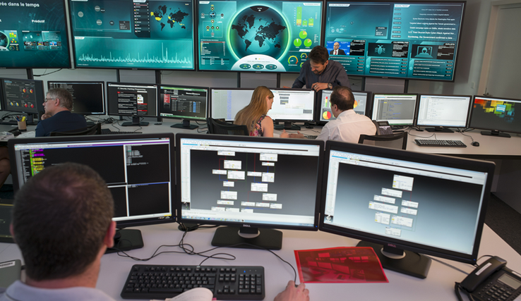 Cinq personnes au travail dans le "Security operation center" de la Direction générale du numérique et des systèmes d'information. On voit de nombreux écrans avec différentes informations. 