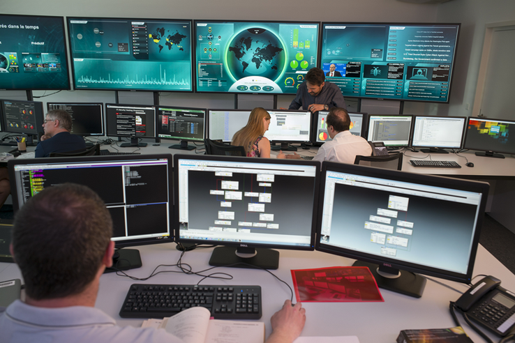Cinq personnes au travail dans le "Security operation center" de la Direction générale du numérique et des systèmes d'information. On voit de nombreux écrans avec différentes informations. 
