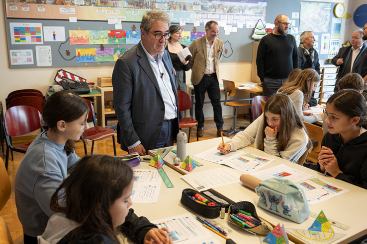 Des élèves autour d'une grand table où sont posés des papiers et des trousses de stylos. Le conseiller d'Etat se tient derrière la table.