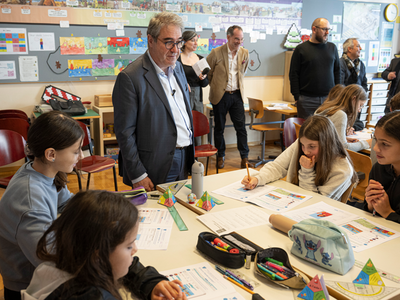 Des élèves autour d'une grand table où sont posés des papiers et des trousses de stylos. Le conseiller d'Etat se tient derrière la table.