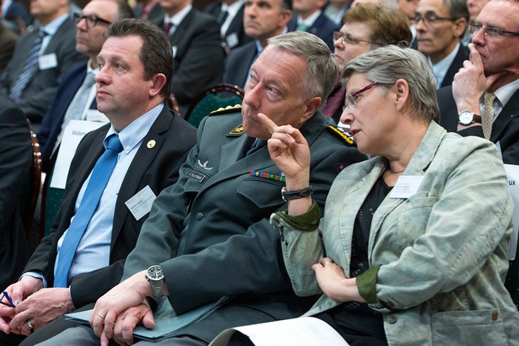 Trois personnages assis dans une salle. Au premier plan Béatrice Métraux désigne quelque chose du doigt à André Blattmann, à sa droite. Le président du Grand Conseil Jacques Nicolet regarde aussi dans la direction pointée.