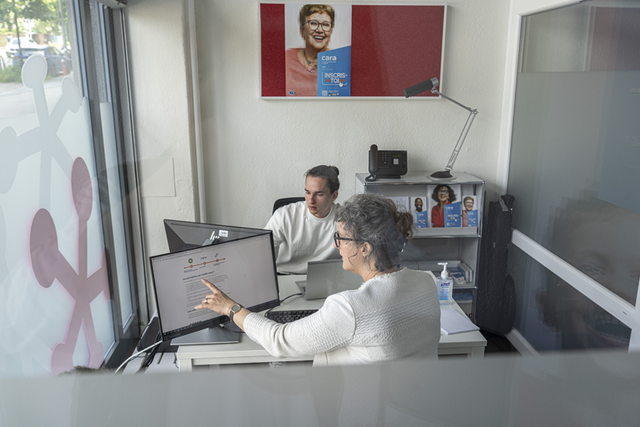 deux personnes face à face dans un petit bureau, chacune avec un ordinateur. L'une explique à l'autre ce qu'il faut faire sur l'ordinateur. Au mur du fond, une affiche sur laquelle on lit: "CARA, inscris-toi".
