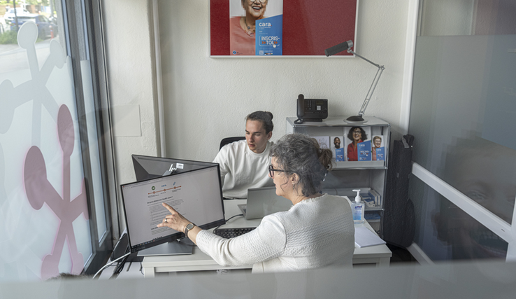 deux personnes face à face dans un petit bureau, chacune avec un ordinateur. L'une explique à l'autre ce qu'il faut faire sur l'ordinateur. Au mur du fond, une affiche sur laquelle on lit: "CARA, inscris-toi".