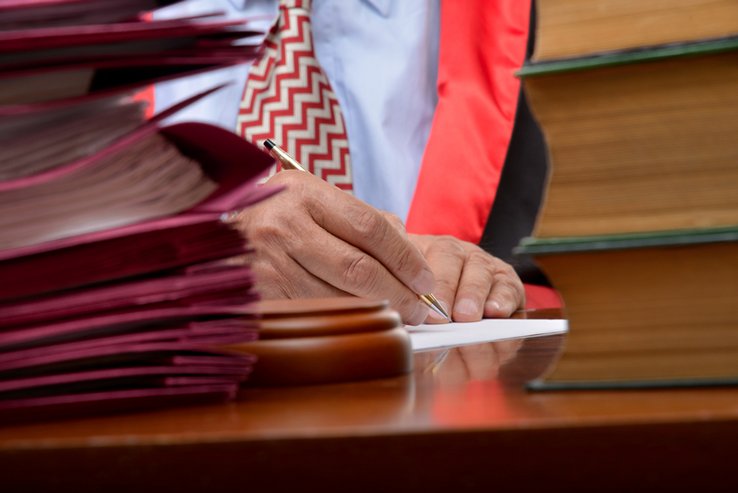 La main d'un juge qui écrit. A côté, une pile de dossiers.