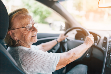 Une dame âgée à son volant, le visage légèrement tourné vers la droite. Elle porte des lunettes et un grand anneau dans l'oreille.