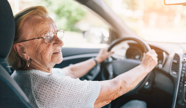 Une dame âgée à son volant, le visage légèrement tourné vers la droite. Elle porte des lunettes et un grand anneau dans l'oreille.