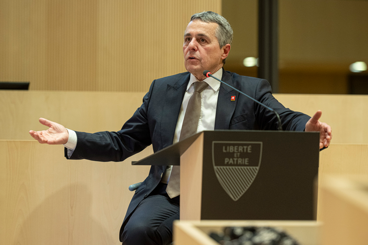 Le conseiller fédéral parle, assis à la tribune, en tendant la main droite.