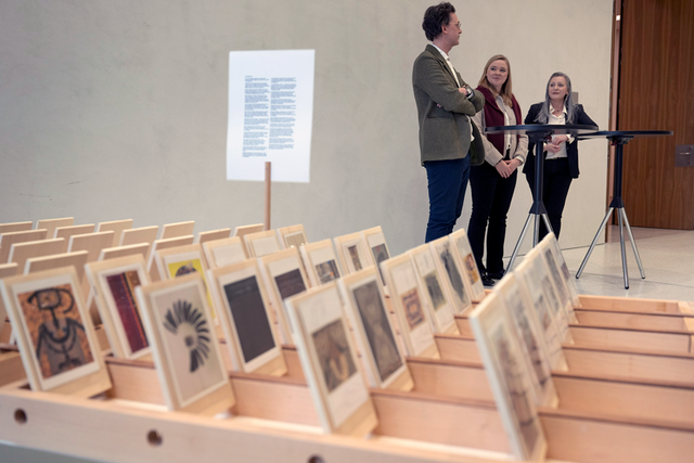 Les trois personnes bavardent au second plan, derrière une série d'images (sorte de cartes postales) des 130 oeuvres exposées.