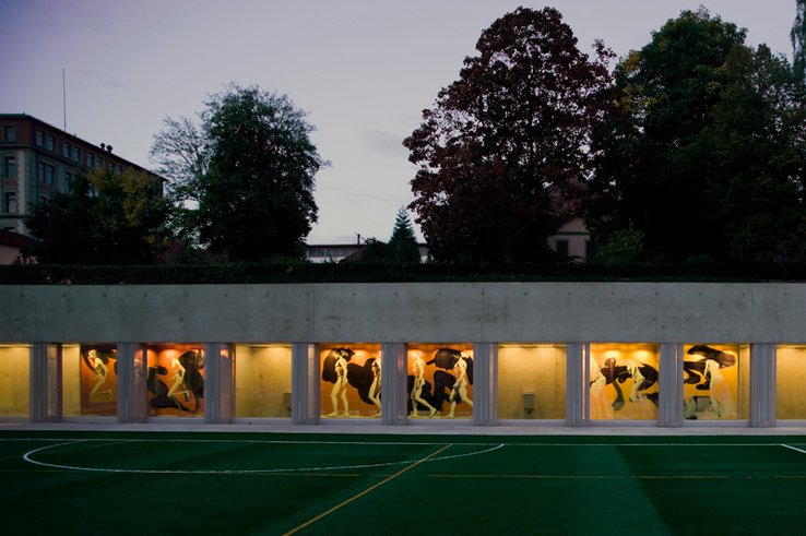 Couloir éclairé avec fresques vues à travers les baies depuis le terrain de sport.