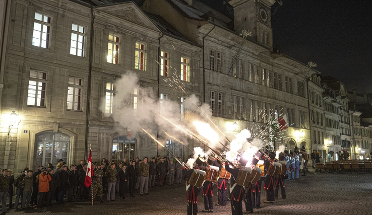 Les Milices vaudoises face à l'Hôtel de Ville de Lausanne, de nuit tirent en l'air avec leurs fusils des cartouches lumineuses. En face, des spectateurs se bouchent les oreilles.