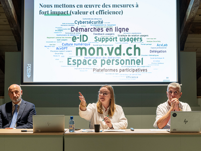 Les personnes sont assises à table durant la conférence de presse. Derrière elles, un grand écran affiche un nuage de mots clés (mon.vd.ch, espace personnel, démarches en ligne...)