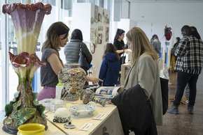 Une salle d'exposition. La restauratrice est derrière une table avec d'anciens vases très décorés. Une visiteuse s'intéresse à une brochure.