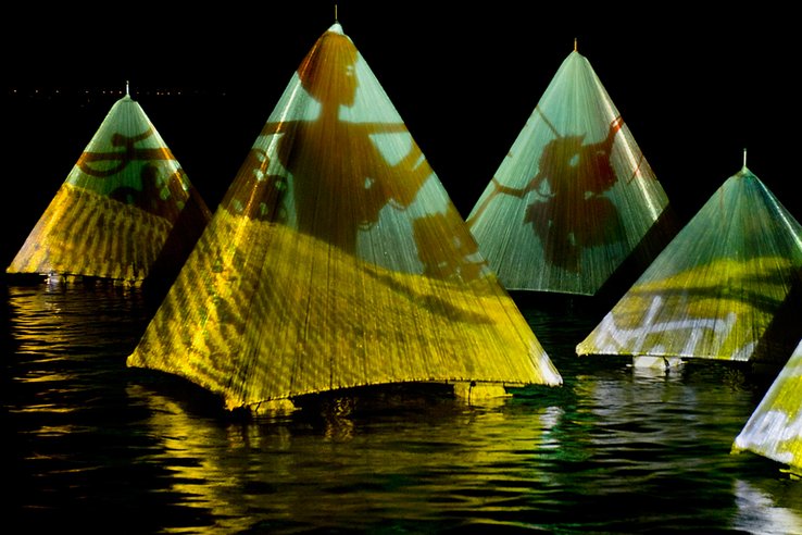 Des pyramides posées au fil de l'eau. En projection sur leurs toiles, des grappes et feuilles de raisin, ainsi qu'une silhouette humaine qui semble travailler la vigne.