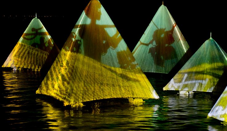 Des pyramides posées au fil de l'eau. En projection sur leurs toiles, des grappes et feuilles de raisin, ainsi qu'une silhouette humaine qui semble travailler la vigne.