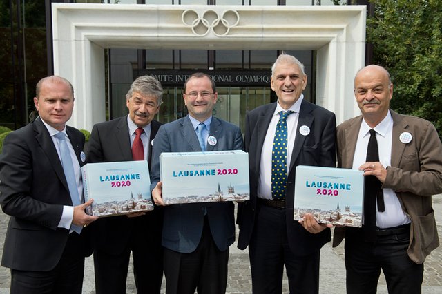 Les personnages posent devant le bâtiment du CIO, tenant en mains trois cartons (le dossier) marqués "Lausanne 2020".
