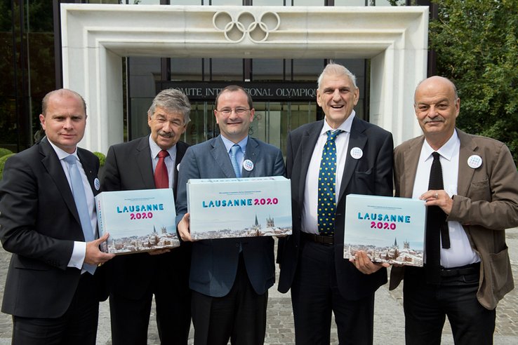 Les personnages posent devant le bâtiment du CIO, tenant en mains trois cartons (le dossier) marqués "Lausanne 2020".
