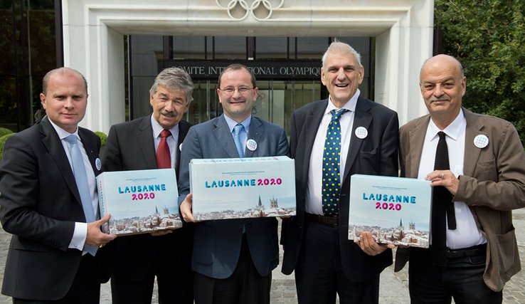 Les personnages posent devant le bâtiment du CIO, tenant en mains trois cartons (le dossier) marqués "Lausanne 2020".