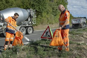 Deux travailleurs au bord de la route ramassent des déchets avec des pinces et un sac.