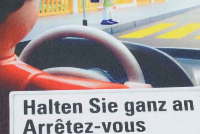 affiche de la campagne: conducteur au volant vu de dos et enfant s'apprêtant à traverser un passage protégé (personnages sous forme de jouets Playmobil)