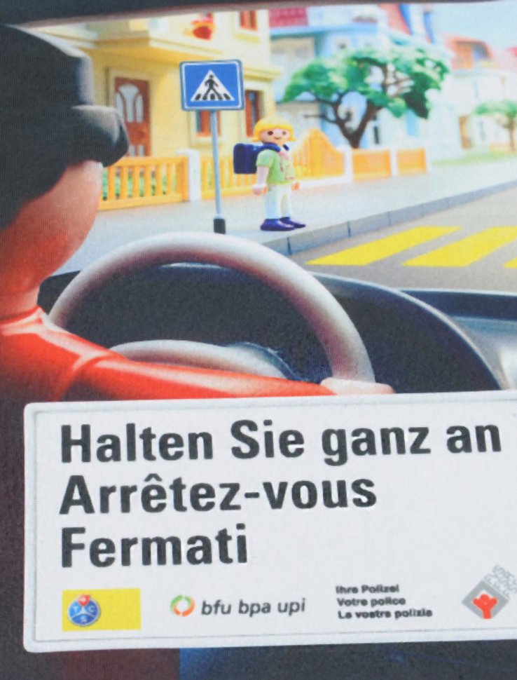 affiche de la campagne: conducteur au volant vu de dos et enfant s'apprêtant à traverser un passage protégé (personnages sous forme de jouets Playmobil)