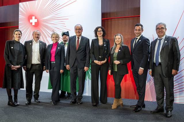 Les personnes posent devant un décor orné d'une croix nationale stylisée.
