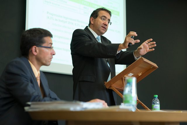 Monsieur Birchmeier est assis et Monsieur Broulis, debout, parle, les deux mains projetées devant lui.