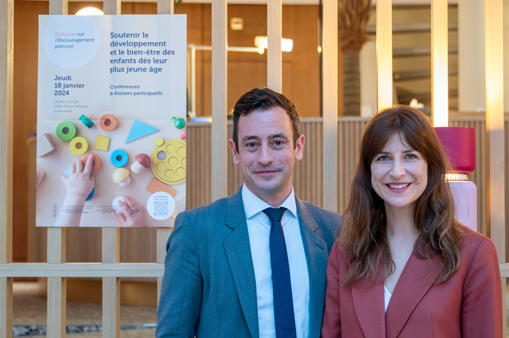 Les deux personnes posent devant une affiche du colloque pour l'encouragement précoce du 18 janvier 2024.