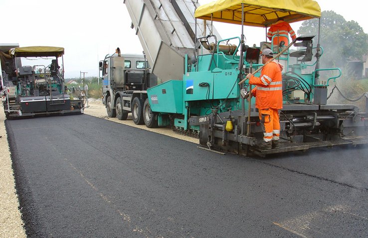 Deux machines posent un revêtement bitumineux. Un homme se tient sur le marchepied d'une goudronneuse.