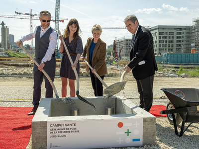 Le quatre personnes tiennent chacune une bêche et versent du ciment par dessus un cylindre d'acier, dans le radier du futur bâtiment. Au fond, des grues et d'autres immeubles en construction, près de la Bourdonnette.