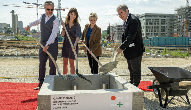 Le quatre personnes tiennent chacune une bêche et versent du ciment par dessus un cylindre d'acier, dans le radier du futur bâtiment. Au fond, des grues et d'autres immeubles en construction, près de la Bourdonnette.