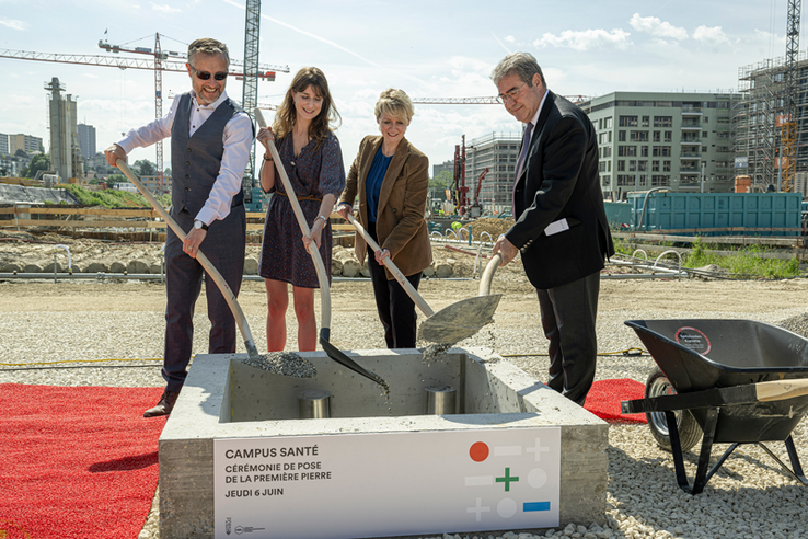 Le quatre personnes tiennent chacune une bêche et versent du ciment par dessus un cylindre d'acier, dans le radier du futur bâtiment. Au fond, des grues et d'autres immeubles en construction, près de la Bourdonnette.