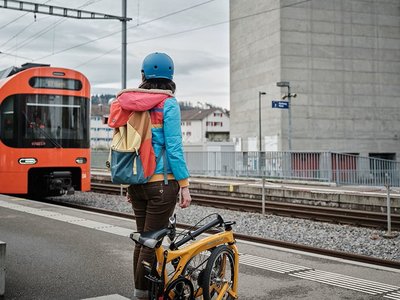 On voit une personne de dos avec un vélo replié sur sa droite, debout sur le quai d'une gare où un train arrive.